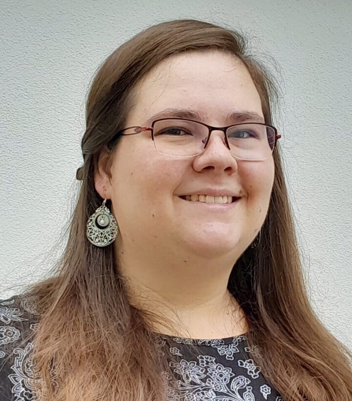 Katharina Hoffmann trägt ein dunkles Oberteil mit filigranem silbernen Muster. Sie hat lange braune Haare, trägt Ohrringe und eine Brille und lächelt.  