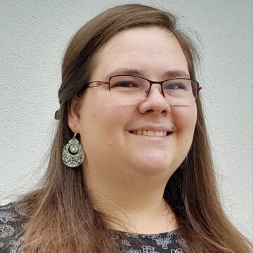 Katharina Hoffmann trägt ein dunkles Oberteil mit filigranem silbernen Muster. Sie hat lange braune Haare, trägt Ohrringe und eine Brille und lächelt.  