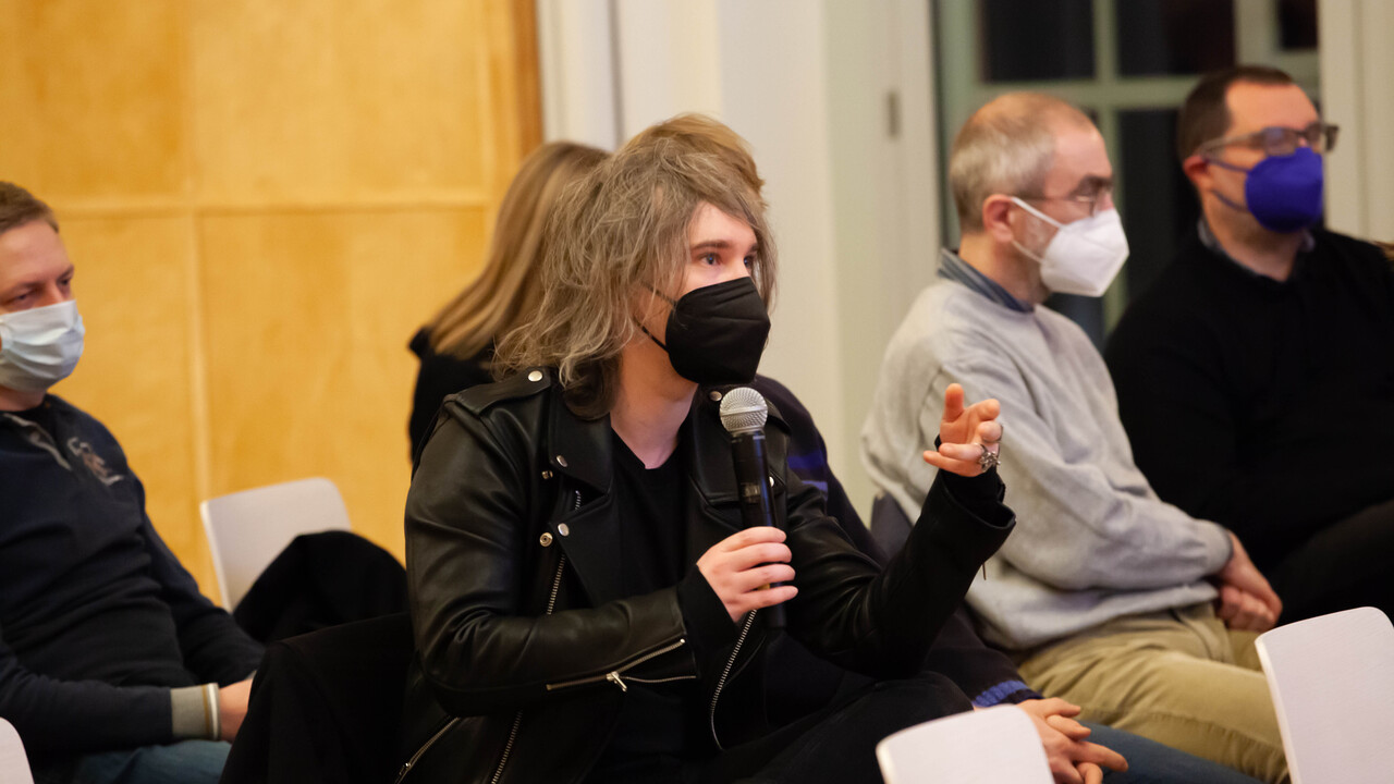 Frage aus dem Publikum: Eine sitzende Person mit schwarzer Corona-Atmschutzmaske hält ein Mikrofon in der rechten Hand in Mundhöhe und blickt mit der linken Hand vor sich gestikulierend rechts aus dem Bild. Um die Person herum sitzen 4 weitere Menschen mit Atemschutzmasken. Den Hintergrund bilden von links nach rechts Holzvertäfelung und weiße Wand mit angeschnittenem Fenster nach außen.