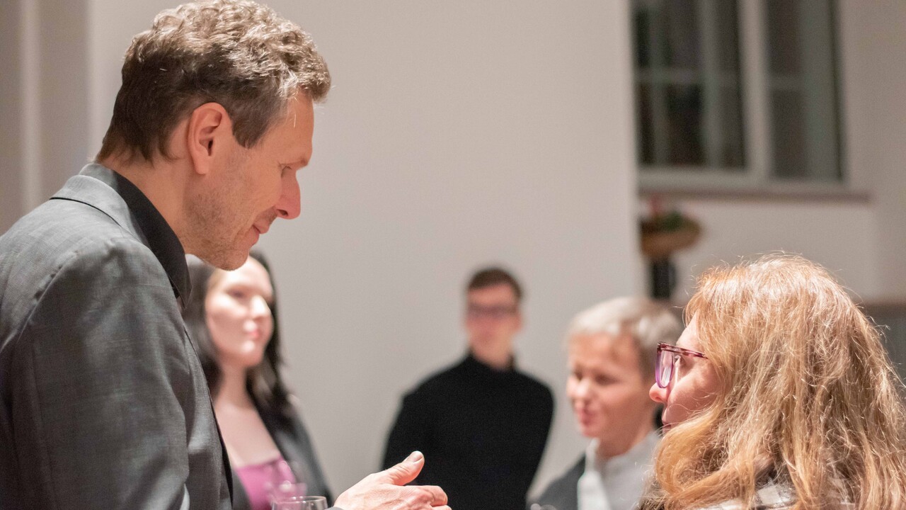 Empfang in den „Katakomben“: Prof. Dr. Tobias Rosefeldt wendet ich einer Frau mit rötlichen Haaren und Brille zu. Von beiden sind die Köpfe von der Seite zu sehen. Im Hintergrund unscharf weitere Personen vor einer weißen Wand mit Fenster.