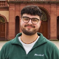 Daniel Wiggenhauser hat braune Haare und einen Vollbart. Er trägt einen grünen Pullover und trägt eine Brille.