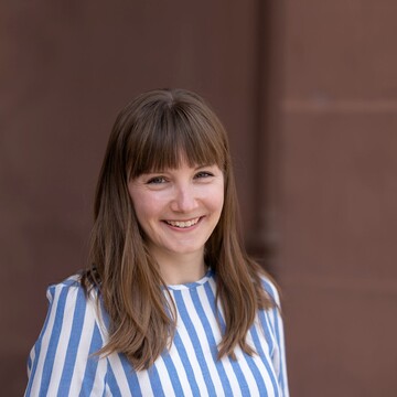 Caterina Katzer hat braune lange Haare und ein Pony. Sie trägt ein blau-weiß-gestreiftes Oberteil.