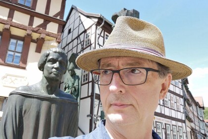 Mann mit Hut und Brille vor einer Statue und Fachwerkhäusern