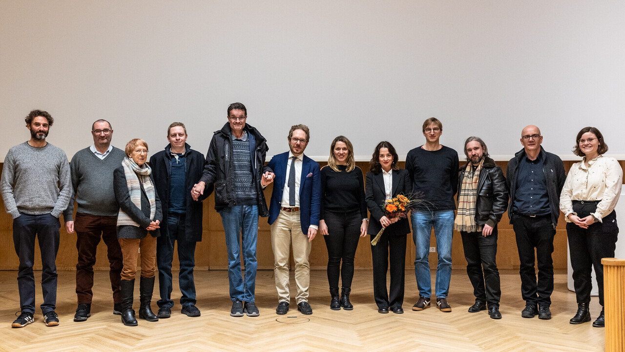 Welttag der Philosophie 2025: Gruppenfoto auf der Bühne Gruppenfoto auf der dem Parkettboden der holzvertäfelten Bühne der Aula der Universität Mannheim zum Tag der Philosophie am 20.11.2025. Mitglieder des Philosophischen Seminars umranden in herbstlicher Kleidung die Vortragende des Abends Elif Özmen, die einen Blumenstrauß in den Händen hält.