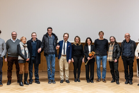 Gruppenfoto auf der dem Parkettboden der holzvertäfelten Bühne der Aula der Universität Mannheim zum Tag der Philosophie am 20.11.2025. Mitglieder des Philosophischen Seminars umranden in herbstlicher Kleidung die Vortragende des Abends Elif Özmen, die einen Blumenstrauß in den Händen hält.