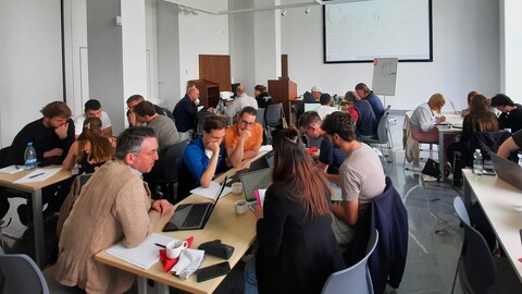 Die Teilnehmenden der Summer School diskutieren in Kleingruppen an Tischen in einem hellen Konferenzraum. Laptops sind aufgeklappt, und eine Präsentation wird an die Wand projiziert.