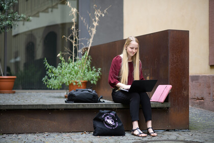 Studentin im Rektoratshof Eine Studentin sitzt mit ihrem Laptop auf dem Schoß draußen und hat Kopfhörer in den Ohren.