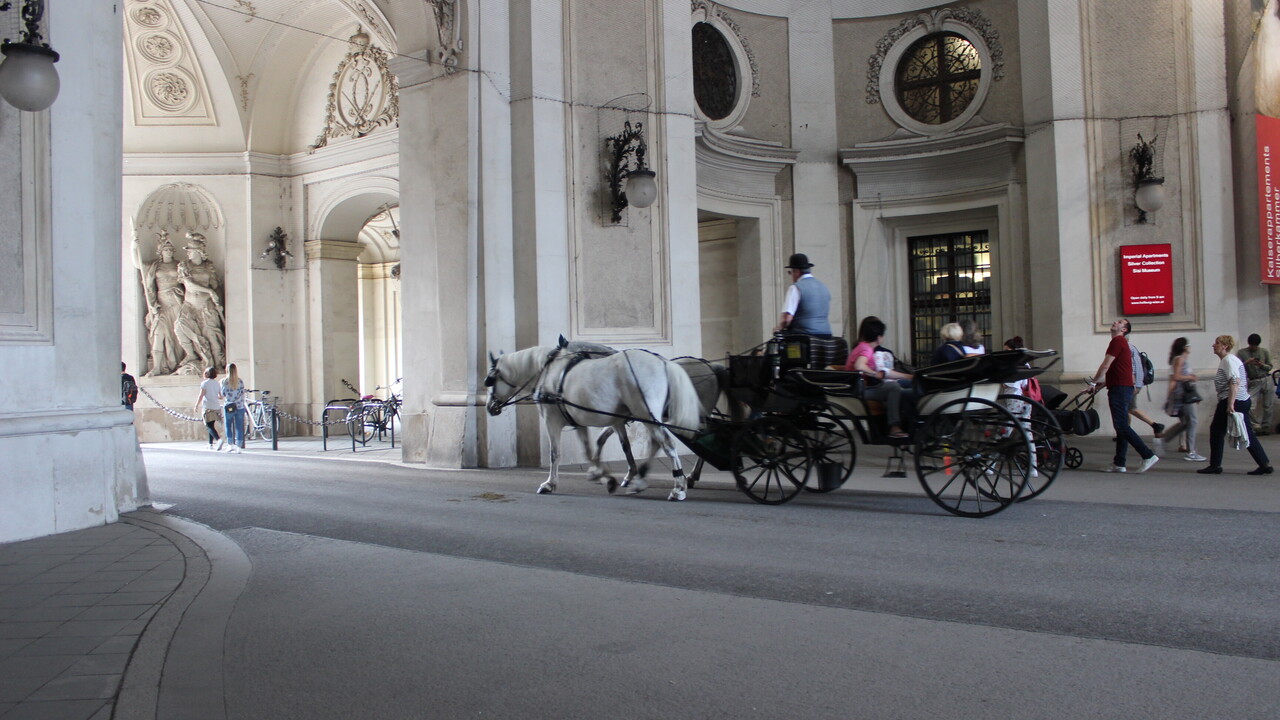 Eine schmale Straße durchquert einen imposanten marmornen Gebäudekomplex. Zentral im Bild ist eine mit 2 Schimmeln bespannte schwarze Kutsche mit Fahrer und Gast. Auf dem dahinterliegenden Gehweg sind diverse Menschen unterwegs.