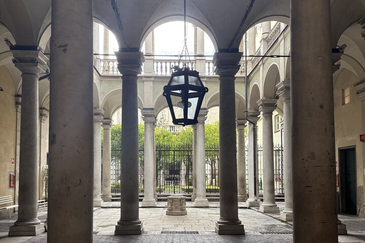 Mittig zu sehen ist eine schwarze Laterne. Dahinter sieht man einen rechteckigen Innenhof, der von einer zweistöckigen Galerie aus beigen Säulen umgeben ist. Im Hintergrund sieht man einen schwarzen Zaun und grüne Büsche.