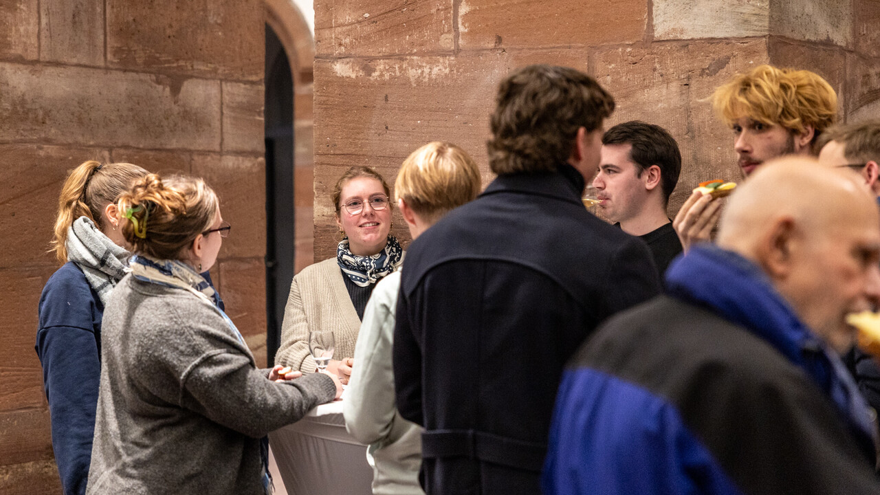 Impressionen vom Empfang in den „Katakomben“ Empfang in den Katakomben der Universität Mannheim am Tag der Philosophie am 20.11.2025: Etwa 10 herbstlich gekleidete Personen mit Gläsern und Snacks in den Händen an Cocktailtischen vor den Säulen aus rotem Sandstein.