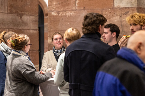 Empfang in den Katakomben der Universität Mannheim am Tag der Philosophie am 20.11.2025: Etwa 10 herbstlich gekleidete Personen mit Gläsern und Snacks in den Händen an Cocktailtischen vor den Säulen aus rotem Sandstein.