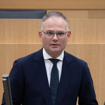 Tim Müller trägt einen schwarzen Anzug und steht an einem Rednerpult. Er hält die Hände vor dem Körper zusammen, trägt eine Brille und hat den Mund leicht geöffnet. 