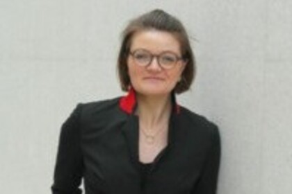 Frau steht vor einer hellen Wand, trägt Brille, schwarzen Blazer mit rotem Kragen und eine Halskette