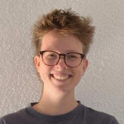 Anne Jaeschke hat kurze blonde Haare und lacht in die Kamera. Sie trägt ein blaues T-Shirt und eine brauen Hornbrille.