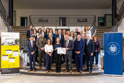 Auf einer Treppe steht eine Gruppe von Personen. In der ersten Reihe stehen eine Frau und ein Mann in der Mitte, die jeweils ein Blatt in der Hand präsentieren.