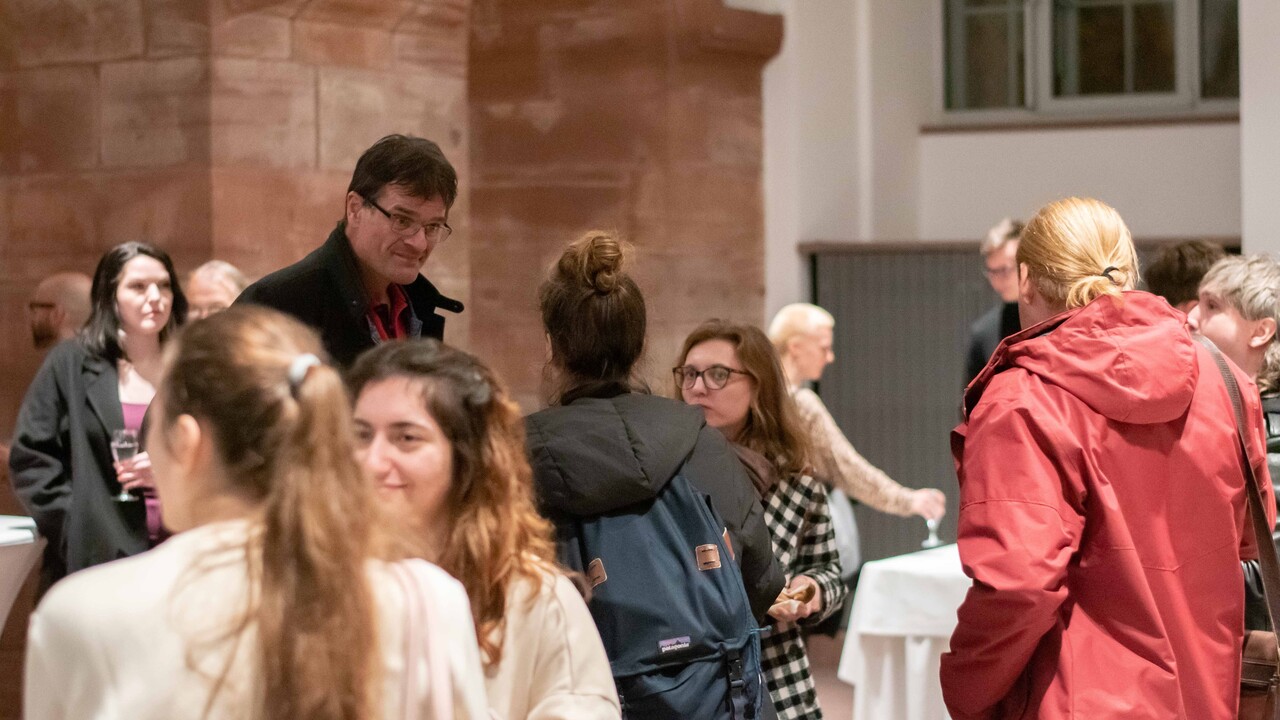 Empfang in den „Katakomben“: Zentral im Bild Prof. Dr. Bernward Gesang im Austausch mit mehreren Gästen. Darum weitere Personengruppen in herbstlicher Kleidung, die teilweise Gläser in der Hand halten. Den Bildhintergrund bilden die roten Sandsteinsbögen und weißen Wände der "Katakomben".