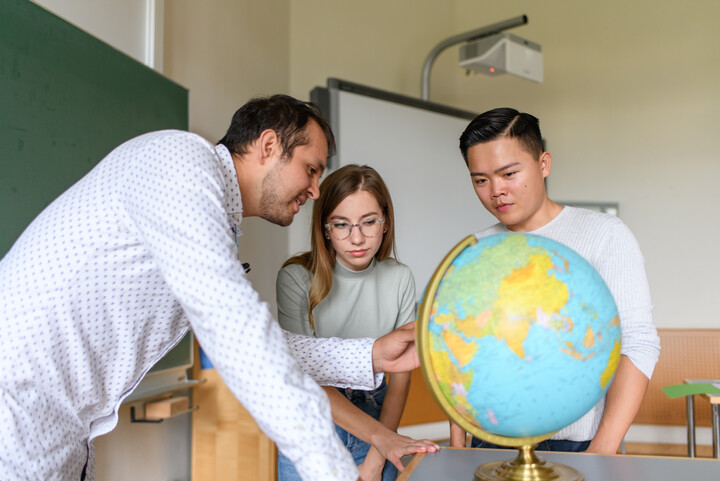 Drei Personen stehen um einen Globus, eine Person zeigt darauf und spricht zu den anderen beiden