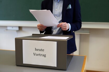 Im Hintergrund eine große grüne Tafel. Davor der mittlere Teil eines Körpers mit hellbelauem Hemd und dunkelblauem Jackett, der mit beiden Händen einen Stapel mit losen Papieren vor sich hält. Kopf und Beine sind nicht zu sehen. Wiederum davor ein Holztisch mit hellgrauer Oberfläche und Rednerpultaufsatz aus dunkelgrau gestrichenen Metall, auf dem weitere Papiere liegen. Auf der Vorderseite des Pultaufsatzes befindet sich ein großer weißer Zettel, auf dem in schwarzer Schrift flächenfüllend "heute Vortrag" gedruckt ist.