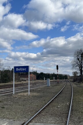 Mehrere Bahngleise, die in der Ferne verschwinden. Links ein paar Gebäude, ansonsten Wald. Im Vordergrund steht ein Schild mit der Aufschrift "Belzec". 