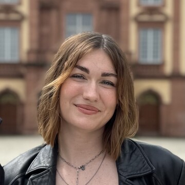 Anna Kuhn vor dem Mannheimer Schloss. Sie trägt braune, schulterlange Haare und eine schwarze Lederjacke.