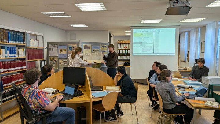 An Tischen in einem Lesesaal sitzende Personen hören zu, wie zwei Personen eine hochgehaltene Urkunde erklären