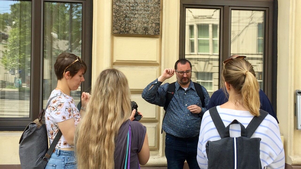 5 Personen in Freizeitkleidung mit Rucksäcken und Taschen vor einer ockerfarbenen Fassade. Zwischen 2 Fenstern ist eine Hinweistafel angebracht, auf der "Kurt Godel" als Überschrift steht. 2 Frauen im Vordergrund stehen mit dem Rücken zur Kamera, eine Frau links ist seitlich zu sehen. Vor der Fassade steht ein Mann in Richtung Kamera und eine weitere Person daneben ist von der Frau links im Vordergrund nahezu komplett verdeckt.