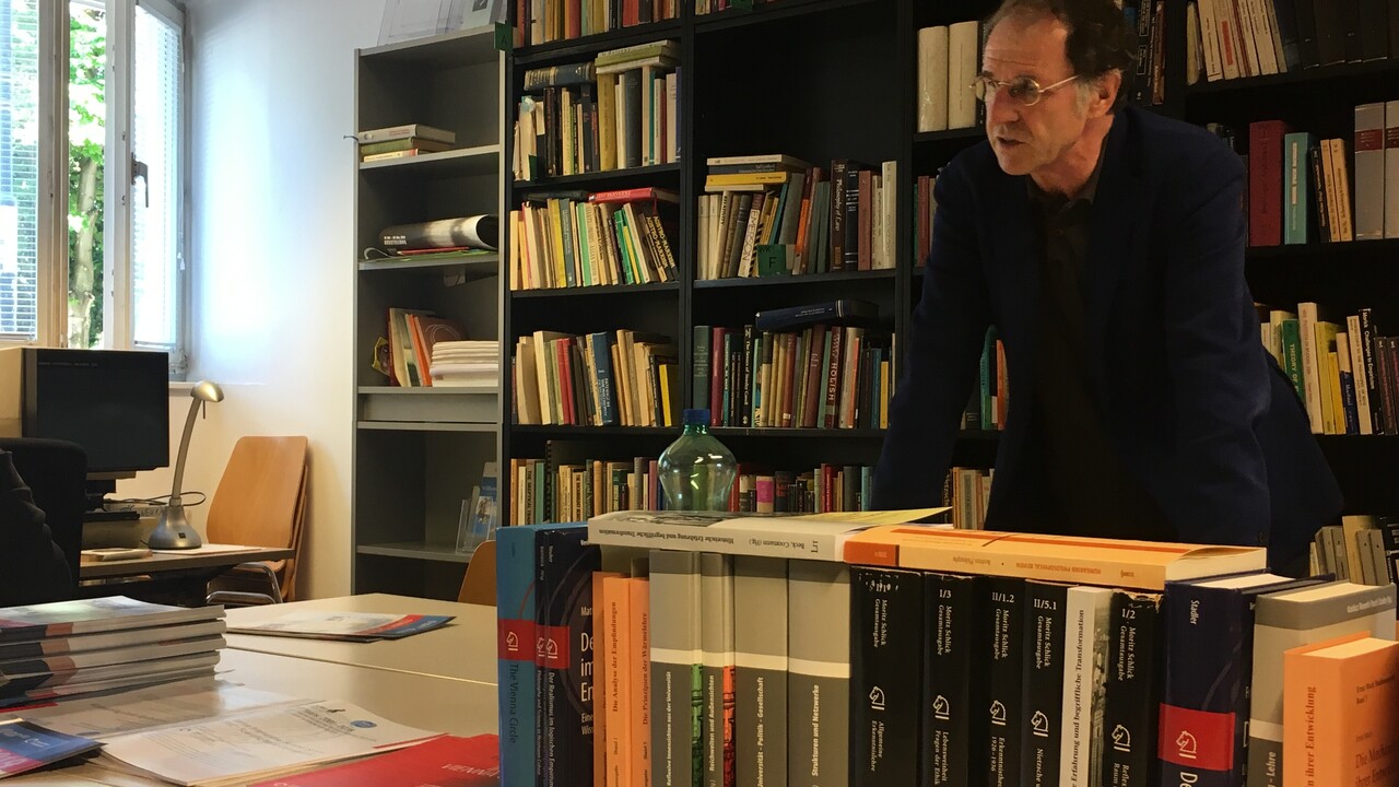 Ein schwarz gekleideter Mann mit Brille stützt sich auf eine Anordnung von Tischen, auf denen viele Bücher und andere Unterlagen liegen und teilweise in einer Reihe stehen. Er steht vor einem schwarzen, gut gefüllten Bücherregal in der rechten Bildhälfte. Links davon schließt sich ein graues Regal an, das neben Büchern auch Flyer und zusammengeklappte Plakate enthält. Ganz links steht ein Betrachtungsgerät - wahrscheinlich für Mikrofiche oder Mikrofilm - auf einem Tisch mit Lampe unter einem halb geöffnetem Fenster.