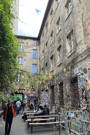 Ein schmaler Hinterhof: Auf der linken Seite wächst Efeu an der Wand entlang. Im Hof stehen mehrere Bierbänke, daran sitzen Menschen. Die alten Steinwände sind bunt bemalt mit Graffitis, Lichterketten hängen über dem Hof. 