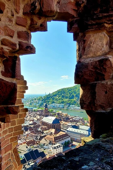 Heidelberger Altstadt mit Stiftskirche und Neckarfluss vom Schloss aus, durch ein ruinöses Fenster hindurch fotografiert.