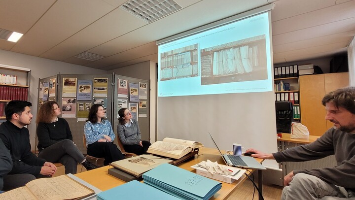 Drei sitzende Frauen und ein Mann schauen auf eine Präsentation auf einer Leinwand, ein Mann an einem Laptop erklärt 