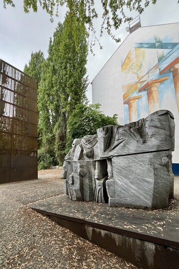 Mahnmal Levetzowstraße: Gedenkort im Freien mit großer rostfarbener Namensstele und fragmentierter Steinskulptur vor einer bemalten Hauswand.