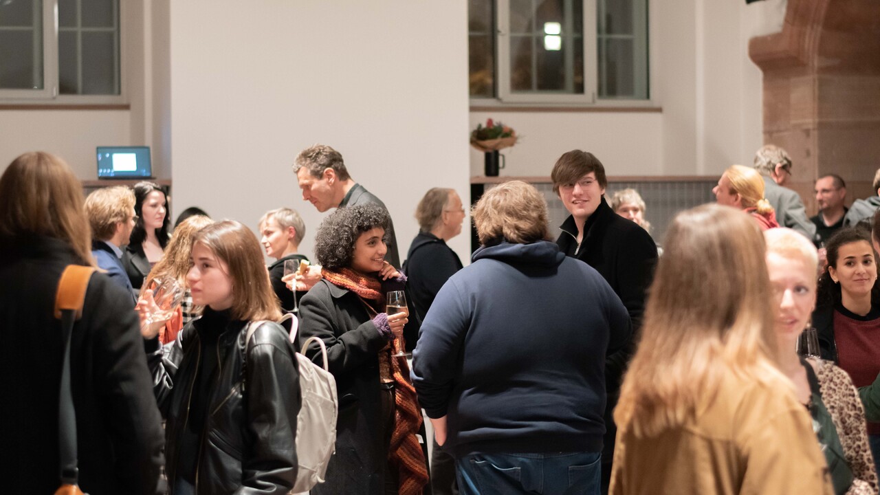 Empfang in den „Katakomben“: Stehende Personengruppen in herbstlicher Kleidung, die teilweise Gläser in der Hand halten, im Gespräch. Im Bildhintergrund eine weiße Wand mit Fenstern, rechts im Anschnitt ein roter Sandsteinsbogen.