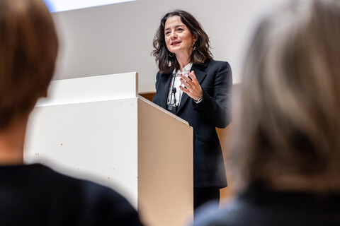 Prof. Dr. phil. Elif Özmen mit umgehängtem Funkmikrofon hinter weißem Rednerpult und gestikulierend gehobener linker Hand. Sie trägt ein helles Hemd und eine dunkle Anzugjacke. Im Hintergrund Leinwand mit angeschnittener Projektion der Bühne in der Aula, im Vordergrund unscharf 2 Hinterköpfe vom sitzenden Saalpublikum.