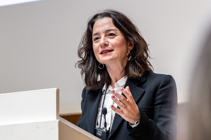 Prof. Dr. phil. Elif Özmen mit umgehängtem Funkmikrofon hinter weißem Rednerpult und gestikulierend gehobener linker Hand beim Vortrag zum Welttag der Philosophie am 20.11.2025. Sie trägt ein helles Hemd und eine dunkle Anzugjacke.