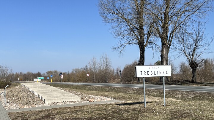 Denkmal für die ehemalige "STACJA TREBLINKA". Auf der einen Seite eine moderne Straße, auf der anderen das Denkmal: Bahngleisen nachempfundene Steinblöcke auf einem Gleisbett aus kleineren Steinen. 