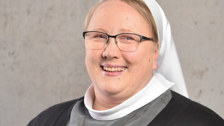 Sr. Marie-Pasquale Reuver trägt ein dunkles Oberteil mit westenähnlichem Ausschnitt, darunter eine weitere Lage grauen Stoff. Um den Hals trägt sie einen weißen Kragen, auf dem Kopf einen weißen Schleier (Wimpel). Über ihrer Brust hängt ein hölzernes Kreuz an einer Kette. Sie trägt eine Brille und lächelt.  