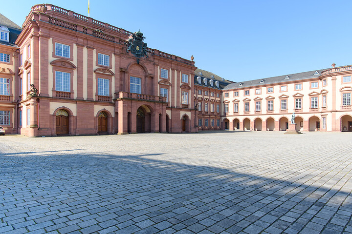Die barocke Fassade des Mannheimer Schlosses im Ehrenhof, mit seinem markanten hellen Sandstein in Gelb und Rot und den symmetrisch angeordneten Fenstern und Bögen unter einem klaren blauen Himmel.