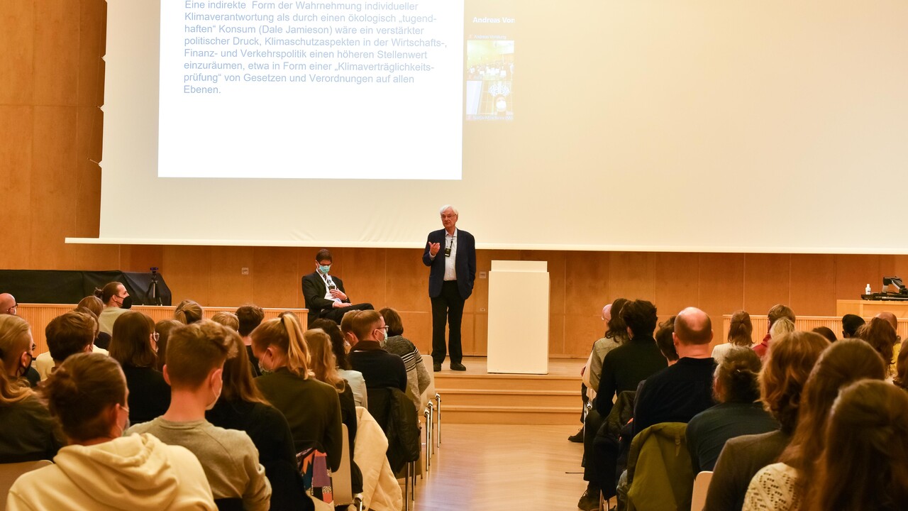 Blick aus dem Mittelganz zwischen dem Publikum auf Prof. Dr. Dr. h.c. Dieter Birnbacher, der auf eine Stelle links im Publikum schaut und gestikuliert. Prof. Birnbacher steht auf der leicht erhöhten Bühne links des Rednerpults, ist bekleidet mit dunkelblauen Anzug und hellblauem Hemd, trägt eine Brille und hat ein Funkmikrofon umgehängt. Etwas links versetzt davon sitzt in schwarzem Anzug, weißem Hemd und in ebenfalls diesen Farben gestreifter Krawatte bei umgehängten Funkmikrofon Prof. Dr. Bernward Gesang. Den Hintergrund des Fotos bildet eine dunkle Holzvertäfelung und helle Projektionsfläche mit angeschnittener projizierter Powerpoint-Folie.