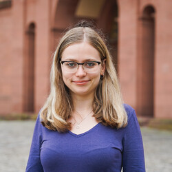Sophia Rishyna in einem lila Oberteil vor dem Mittelbau des Schlosses der Universität Mannheim. Sie trägt langes blondes Haar und eine Brille. 