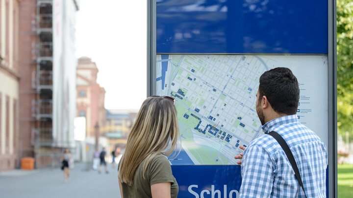 Campusplan Zwei Personen stehen vor einer Infotafel mit einer Karte der verschiedenen Unigebäude.