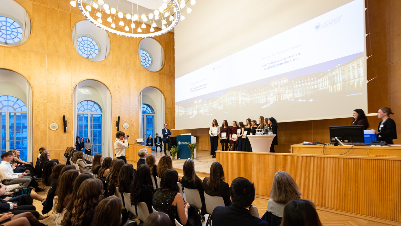 Auf der Bühne der Aula stehen neun Absolvent:innen. Sie halten Ihre Schmuckurkunden in der Hand.