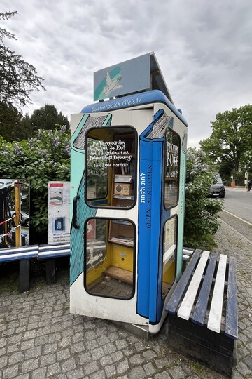 Die Bücherboxx am Mahnmal Gleis 17: Eine umgebaute Telefonzelle, in deren Inneren mehrere Regale übereinander angebracht sind, auf denen Bücher stehen. Auf dem Dach befindet sich ein Solarpanel, die Telefonzelle ist bunt bemalt und beschrieben. 