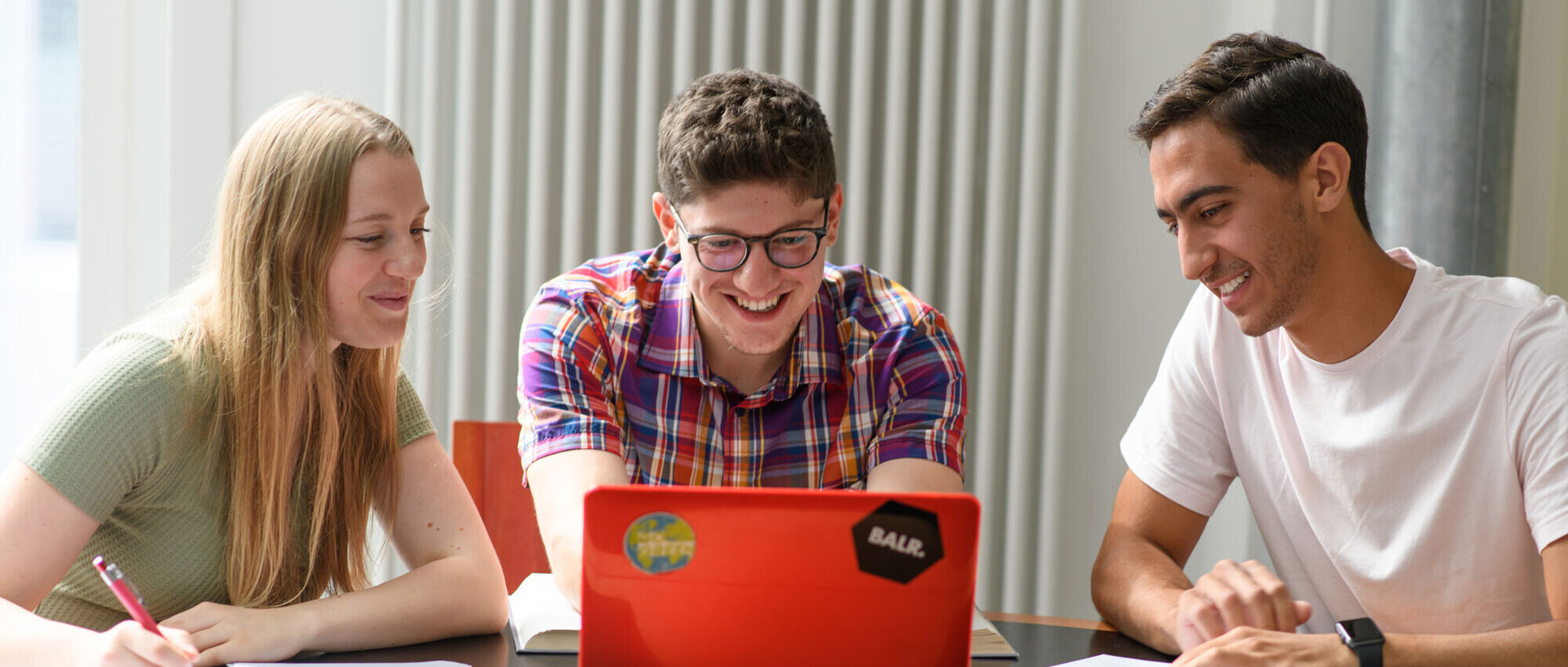 Drei Studierende sitzen mit ihren Arbeitsmaterialien vor einem roten Laptop