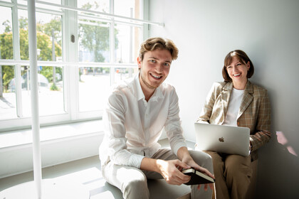 Zwei Forschende sitzen auf einer Fensterbank. Die Frau arbeitet an einem Laptop. Der Mann hält ein Notizbuch in den Händen. Beide lachen. 
