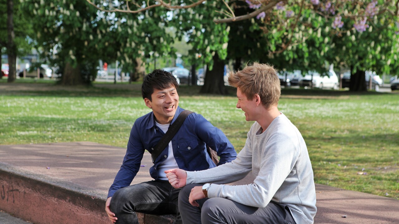 Zwei Studenten sitzen auf Steinen und lachen gemeinsam. Im Hintergrund blühen ein paar Bäume.