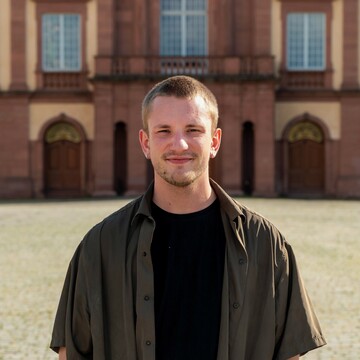 Julian Schmiedel vor dem Mittelbau des Schlosses der Universität Mannheim. Er trägt ein schwarzes Shirt und ein khakifarbenes offenes Hemd darüber. Er hat kurzes blondes Haar, trägt einen Bart und silberne Ohrringe.  