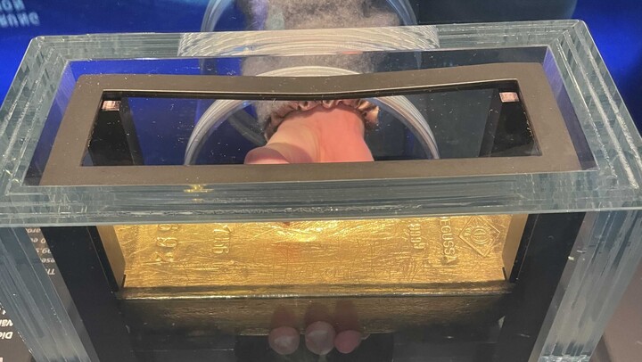 Eine Hand versucht einen Goldbarren in einer Vitrine anzuheben