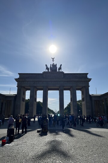 Das Brandenburger Tor aus einiger Entfernung, vor blauem Himmel, die Sonne steht direkt mittig hinter/über dem Tor.