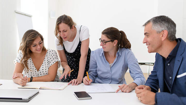 Zwei Forscherinnen und ein Forscher sitzen am Tisch, während eine Forscherin stehend etwas in einem Notizbuch erklärt.