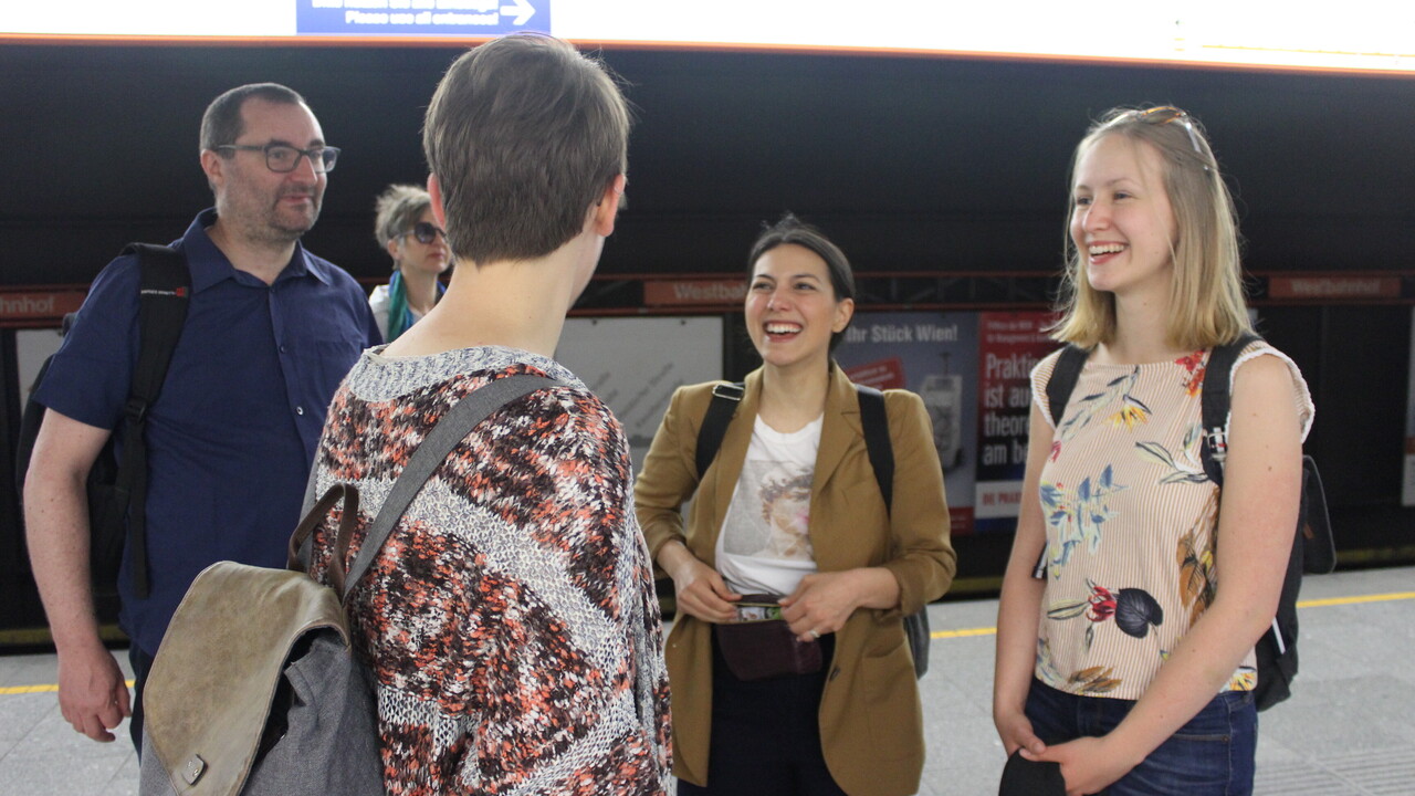 5 Personen in Freizeitkleidung mit Rucksäcken auf einem Bahnsteig mit Plakatwerbung und Haletestellenname "Westbahnhof" im Hintergrund. Die mittlere, etwas im Vordergrund stehende Person dreht der Kamera den Rücken zu. Links davon schaut ein Mann mit normaler Brille lächelnd, etwas abgesetzt eine Frau mit Sonnenbrille interessiert zur Kamera. Rechts stehen zwei lachende junge Frauen.